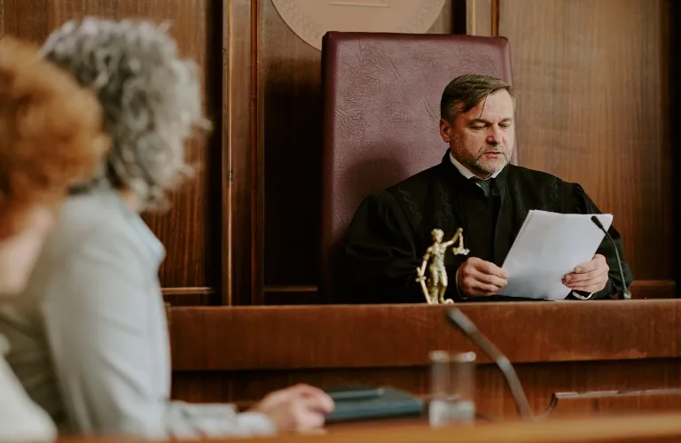 Judge in black robe reading documents in a courtroom with wooden paneling.